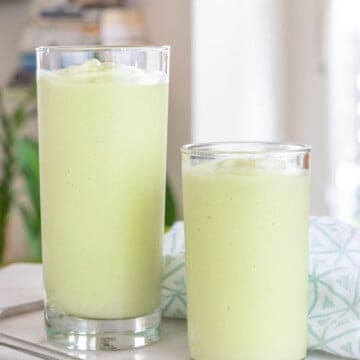 two glasses of completed avocado shakes on a white surface with a metal straw in the background