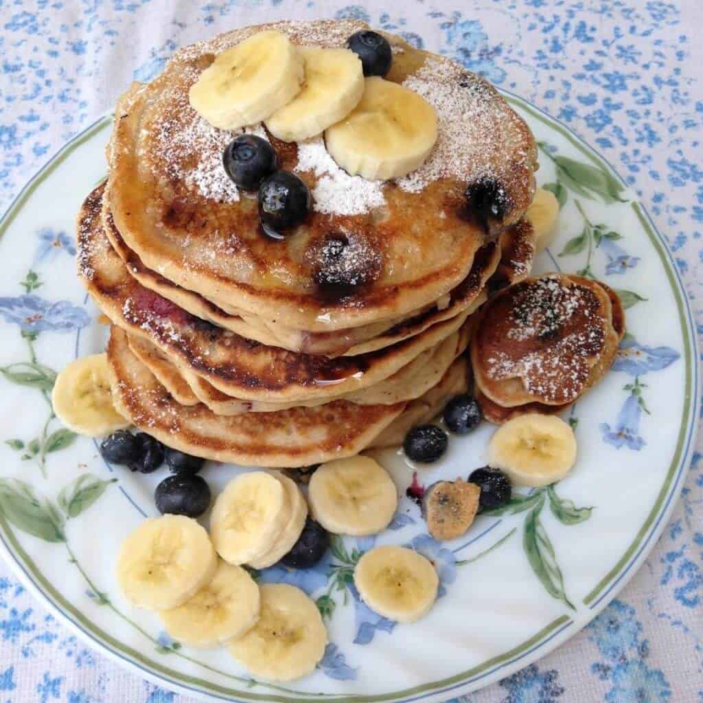 A tall stack of blueberry banana pancakes on a plate with fresh blueberries and banana slices.