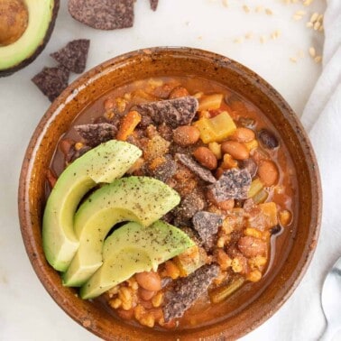 Barley and bean soup in a brown bowl topped with avocado and tortilla chips