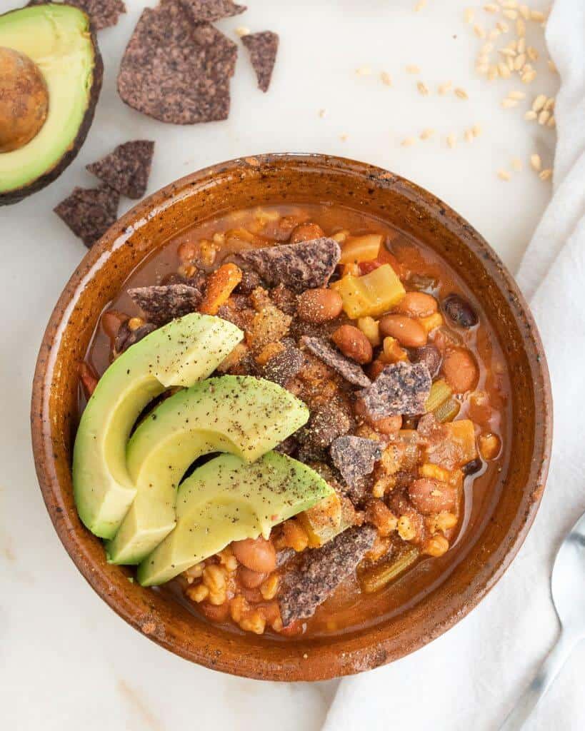 Barley and bean soup in a brown bowl topped with avocado and tortilla chips