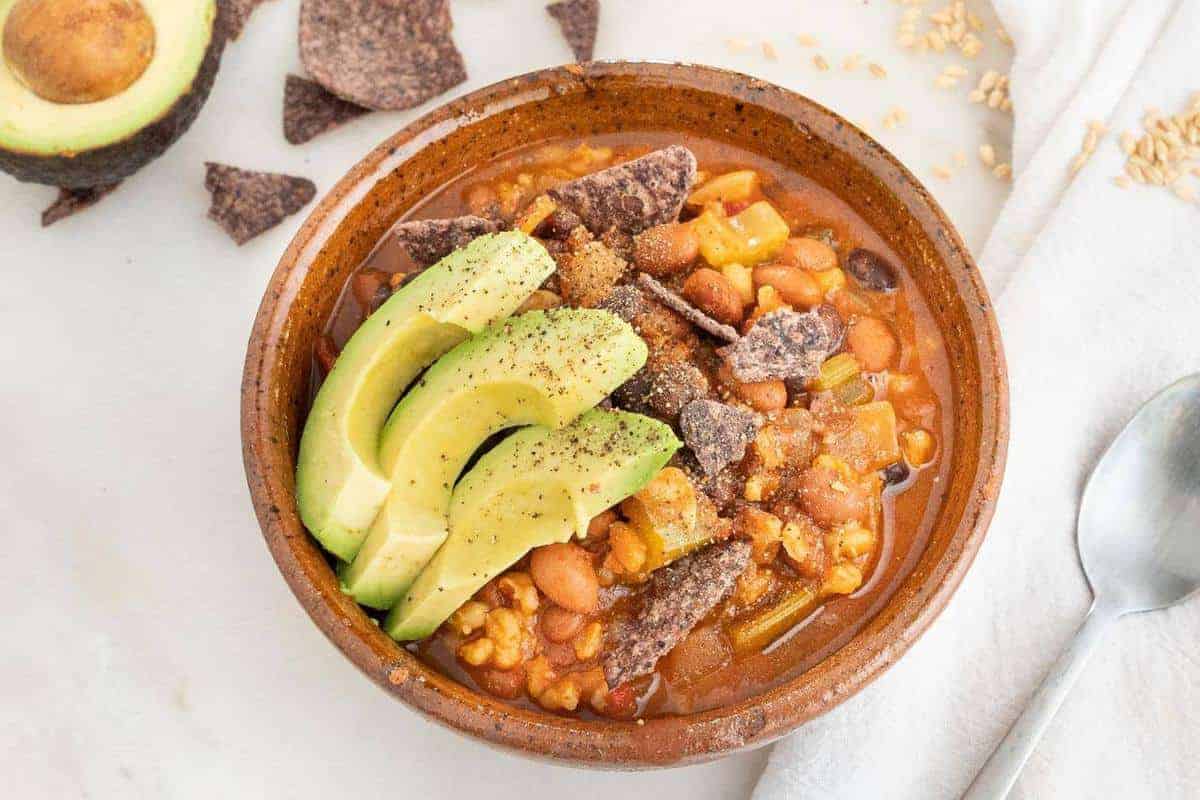 Closeup of bean and barely soup topped with blue corn chips and sliced avocado.