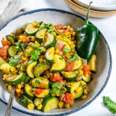 completed calabacitas in a white bowl against a white background with scattered green veggies