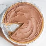 Vegan chocolate pie with one slice cut out. It's in an aluminum pie tin, on top of white and wood cutting board.