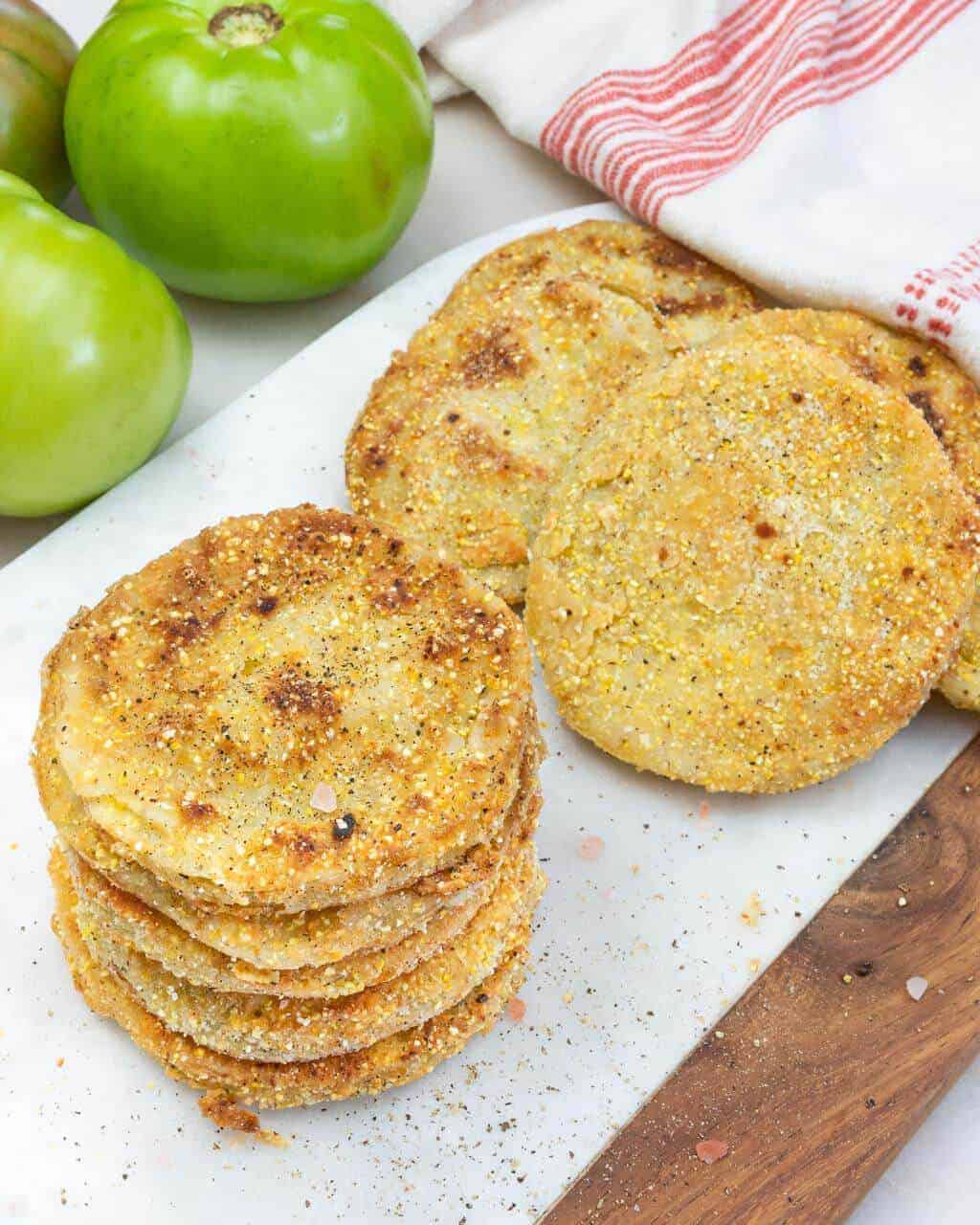 stack of fried green tomatoes on a white background with green whole tomatoes in the background