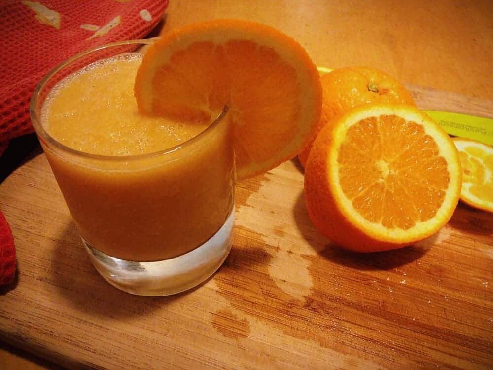 Small glass of orange julius next to a cut orange on a wood counter.