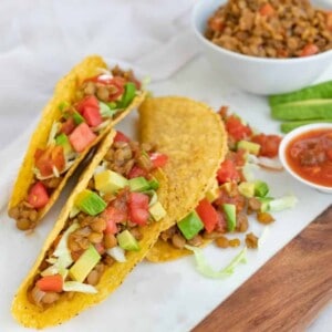 three completed lentil tacos on a white surface with ingredients in the background