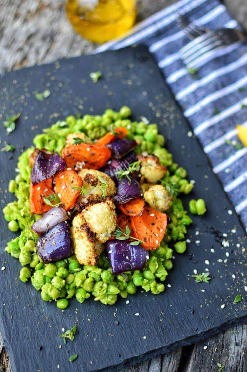 A pile of mushy peas on a black slate topped with roasted veggies.