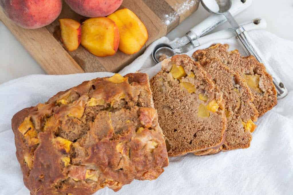 Sliced loaf of peach banana bread on a white cloth with some sliced peaches in the background