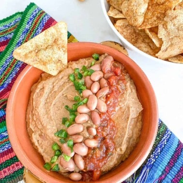 completed pinto dip in an orange bowl against a multi color cloth with a tortilla chip and bean dressing on top