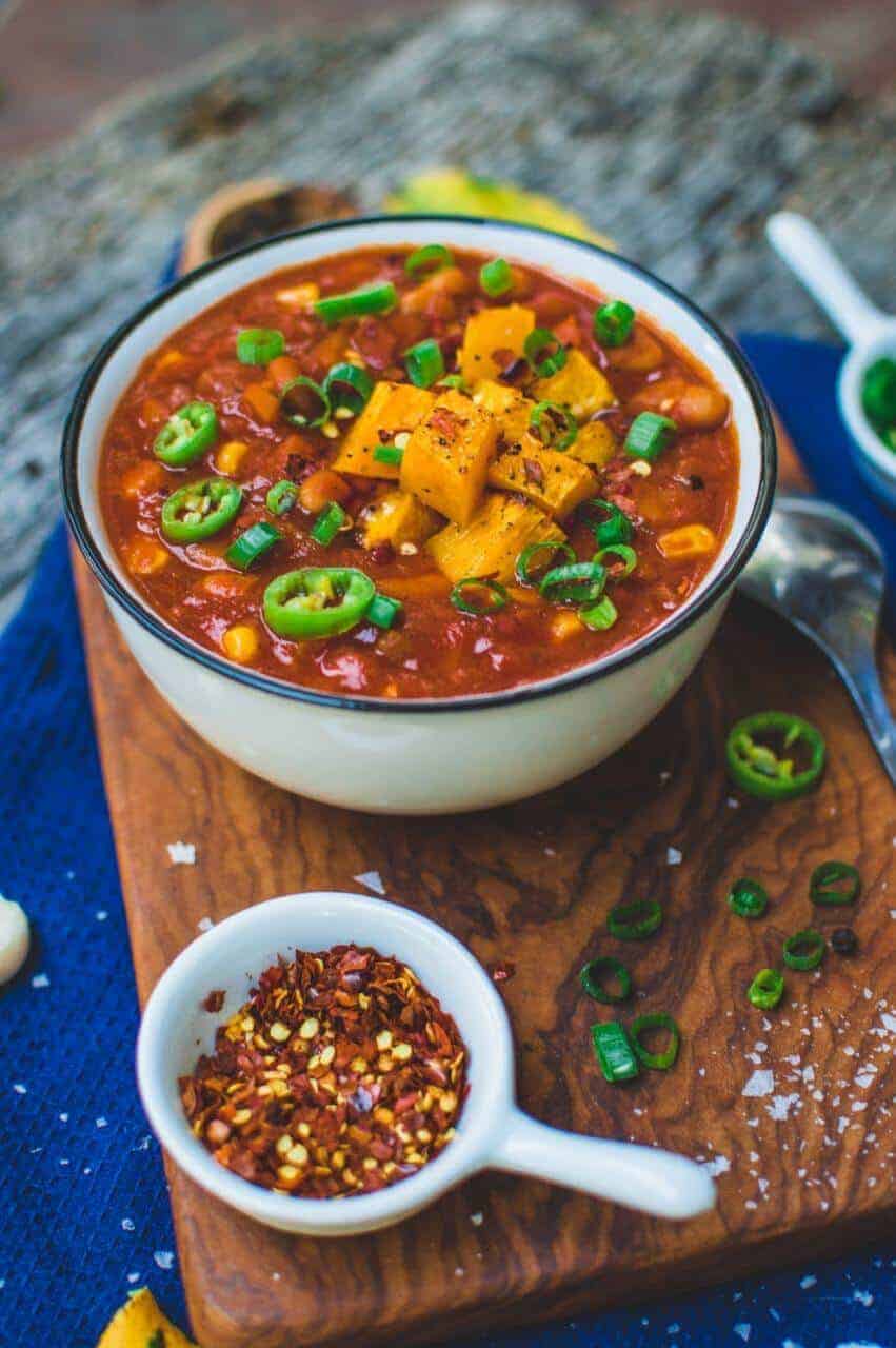 A soup bowl of pumpkin chili on a wood platter next to a pinch bowl of red pepper flakes.