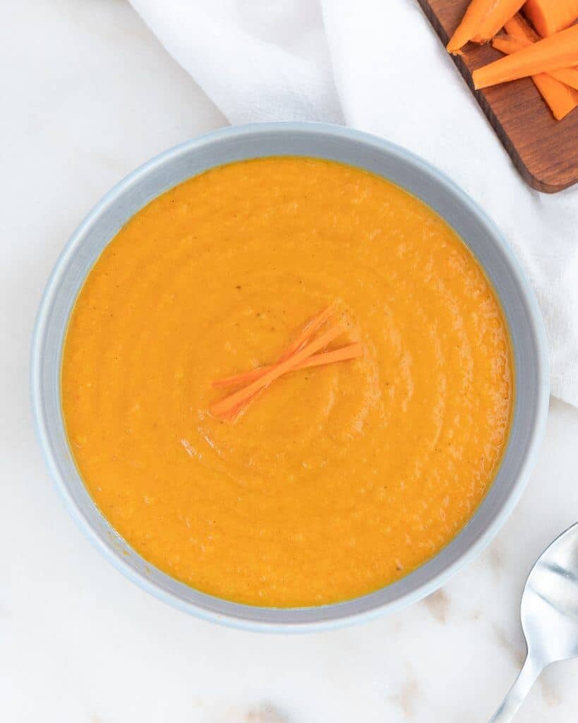 completed spicy carrot soup in blue bowl against white background with cutting board in the background