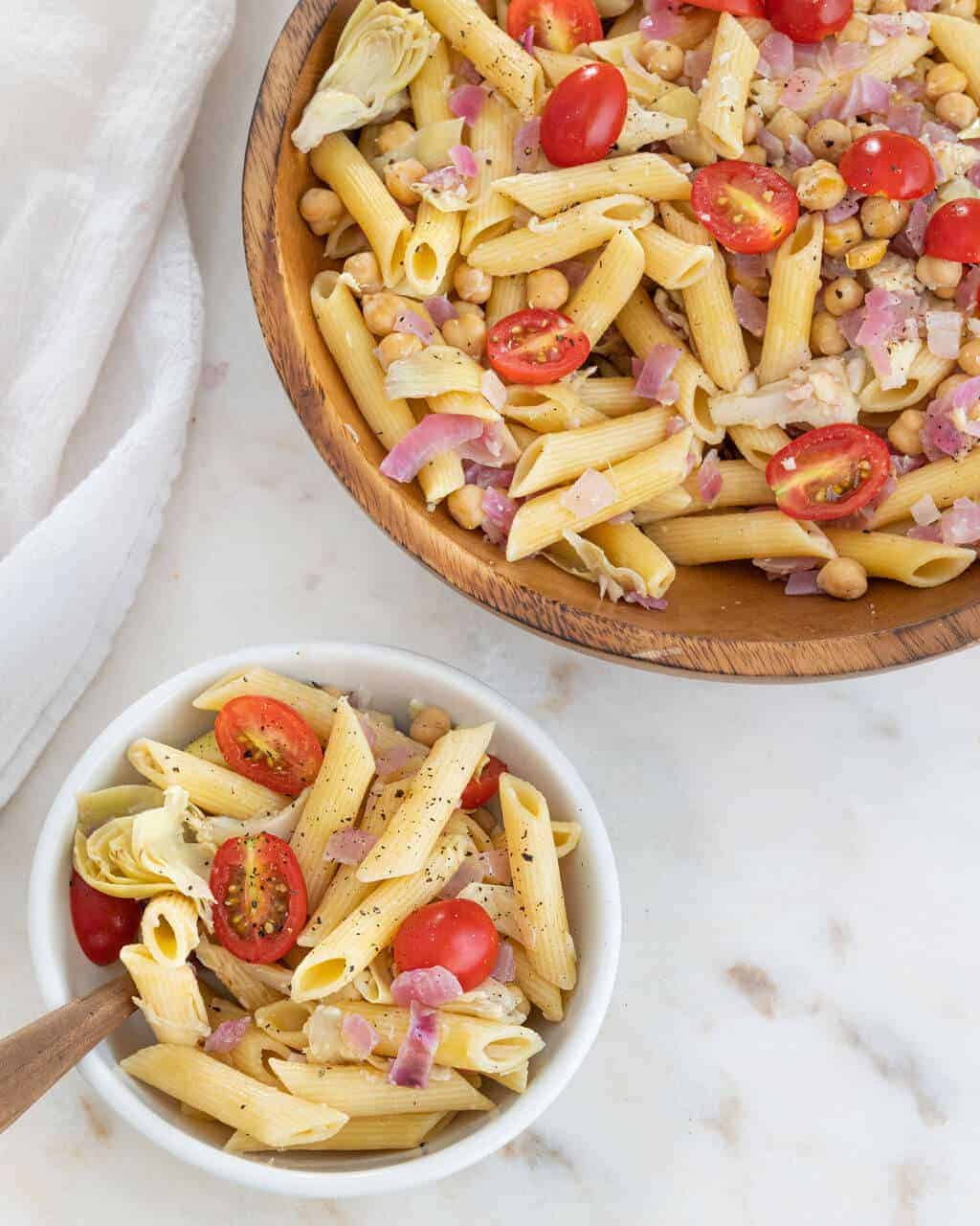 Summer penne and tomato pasta salad in a small bowl next to a full wood serving bowl.
