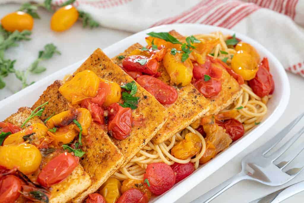 Tofu filets over spaghetti and cherry tomatoes in a long white dish.
