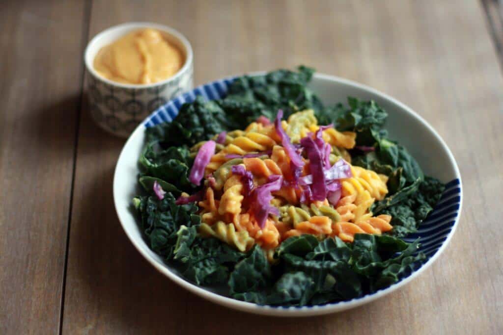 Small cup of vegan cheese sauce next to a bowl of noodles and kale.