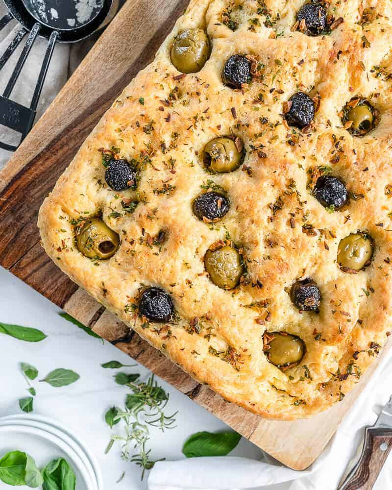 cooked Focaccia bread on cutting board with herbs in the background