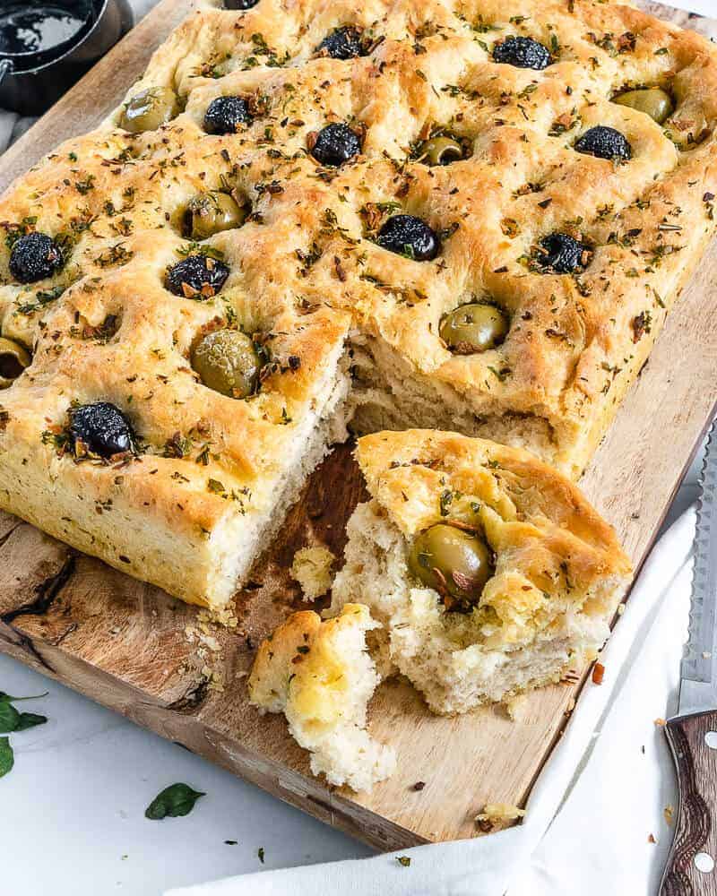 cooked Focaccia Bread with black and green olives with one slice cut on cutting board