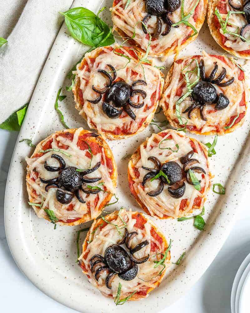 several cooked halloween spider bagel bites on a white platter with ingredients in the background