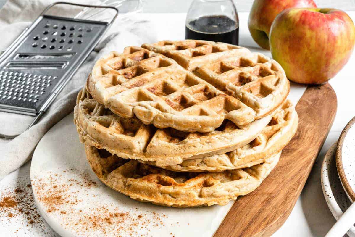 a stack of finished Waffles Apple Cinnamon pancakes on a cutting board with ingredients in the background