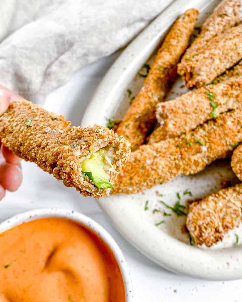 completed Vegan Baked Zucchini Fries on a white platter with one close up fry and a sauce against a white background