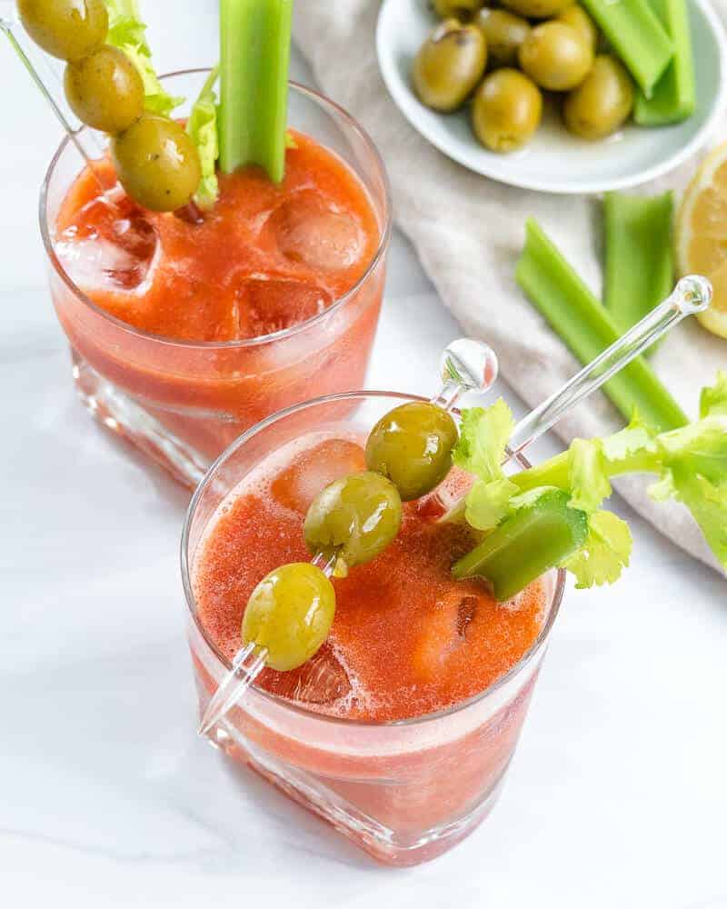 2 Virgin Bloody Mary drinks in glass with decorative olives and veggies in the cup and on the side with a white background