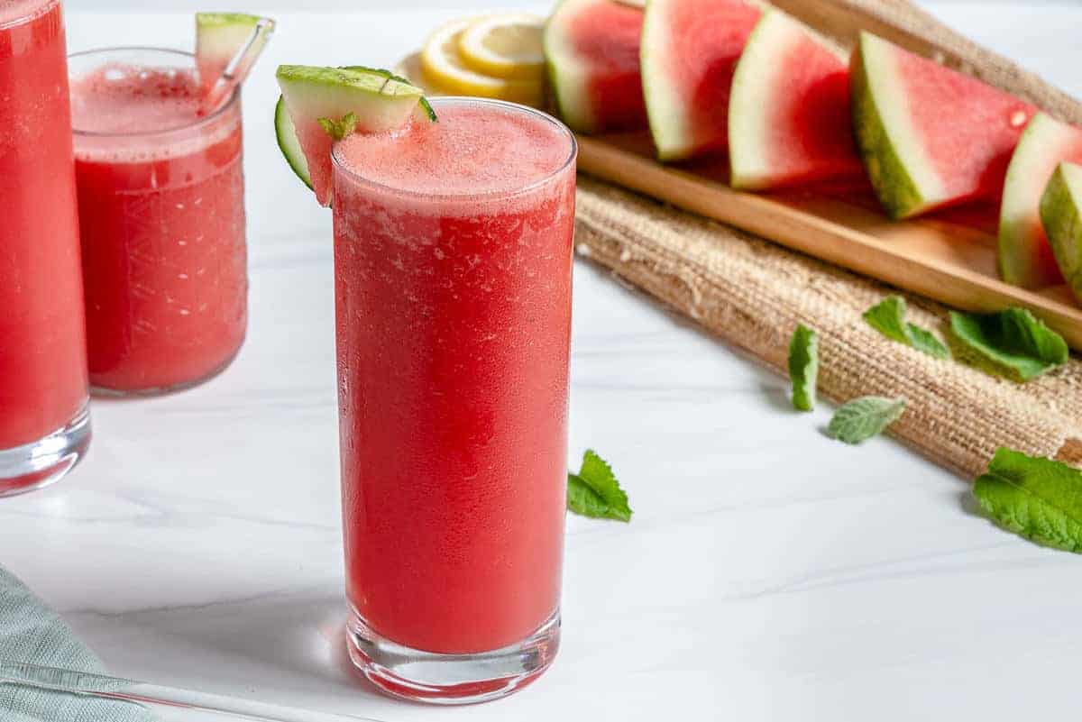 watermelon cooler in tall glass cup with sliced watermelon and more cups of watermelon cooler in a white background