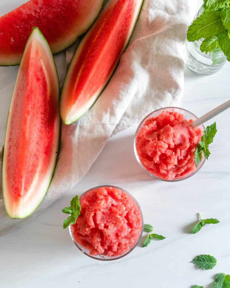 watermelon slushies in two glasses with watermelon wedges in the background.