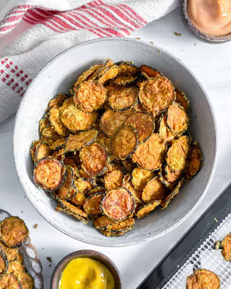 cooked zucchini chip sin a white bowl with zucchini chips in the background