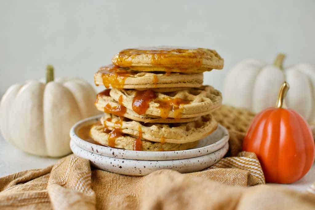 completed Vegan Pumpkin Waffles stacked on top of one another on a white plate with pumpkins in the background