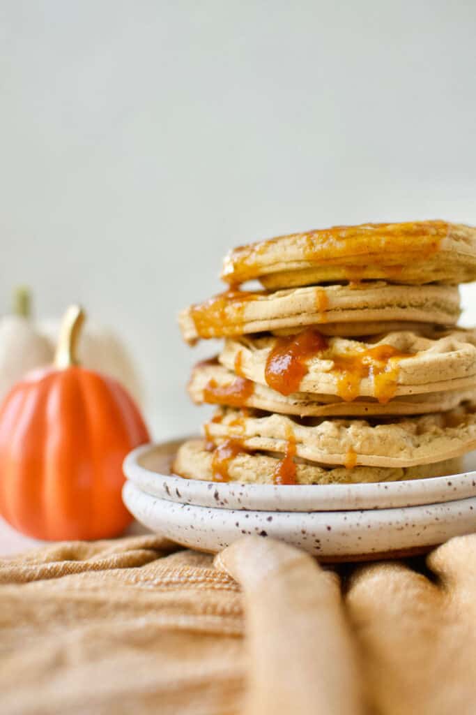 completed Vegan Pumpkin Waffles stacked on top of one another on a white plate with a pumpkin in the background