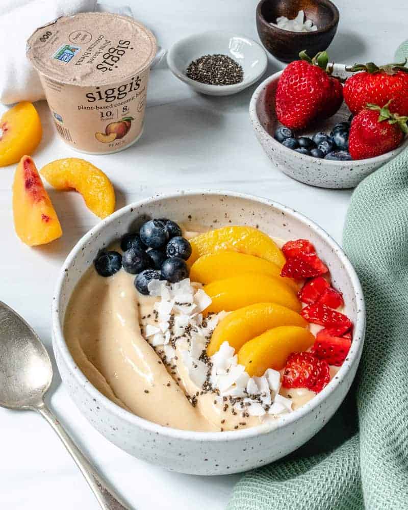 finished smoothie bowl topped with fruit in white bowl with yogurt and fruits scattered against a white background