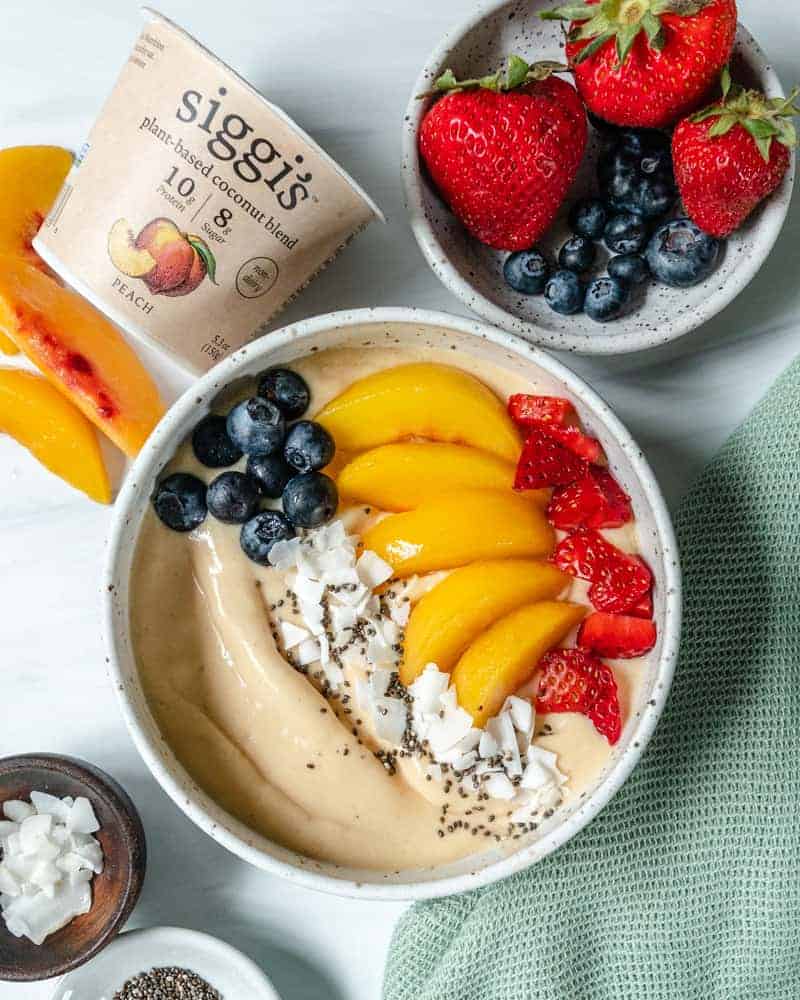 finished smoothie bowl topped with fruit in white bowl with yogurt and fruits scattered against a white background