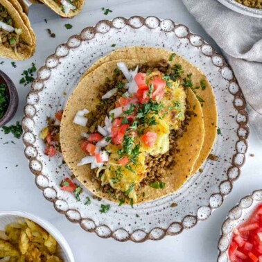 Breakfast Tacos in a white plate with tacos and ingredients in the background against white surface