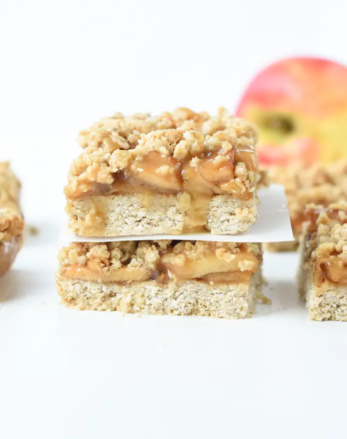 finished vegan apple bars stacked on each other with apples against a white background