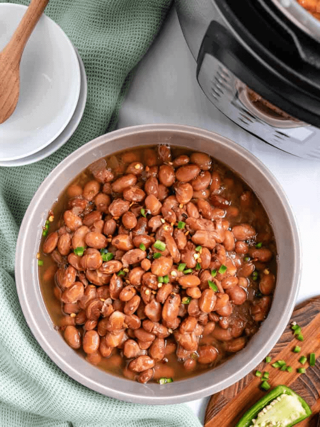 Ready pinto beans in a bowl in front of a white counter with a light green cloth and a brown cutting board in the background