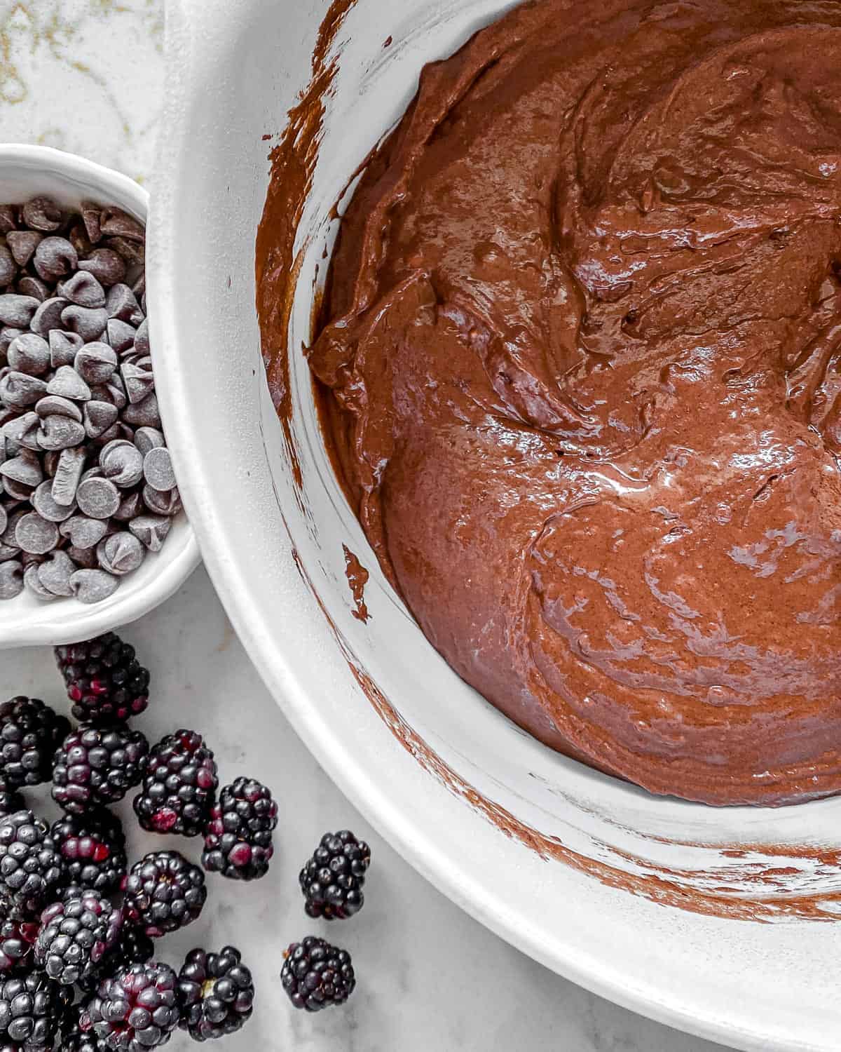 post mixing of wet and dry ingredients for Blackberry Chocolate Cupcakes in white bowl with chocolate chips and blackberries in the background