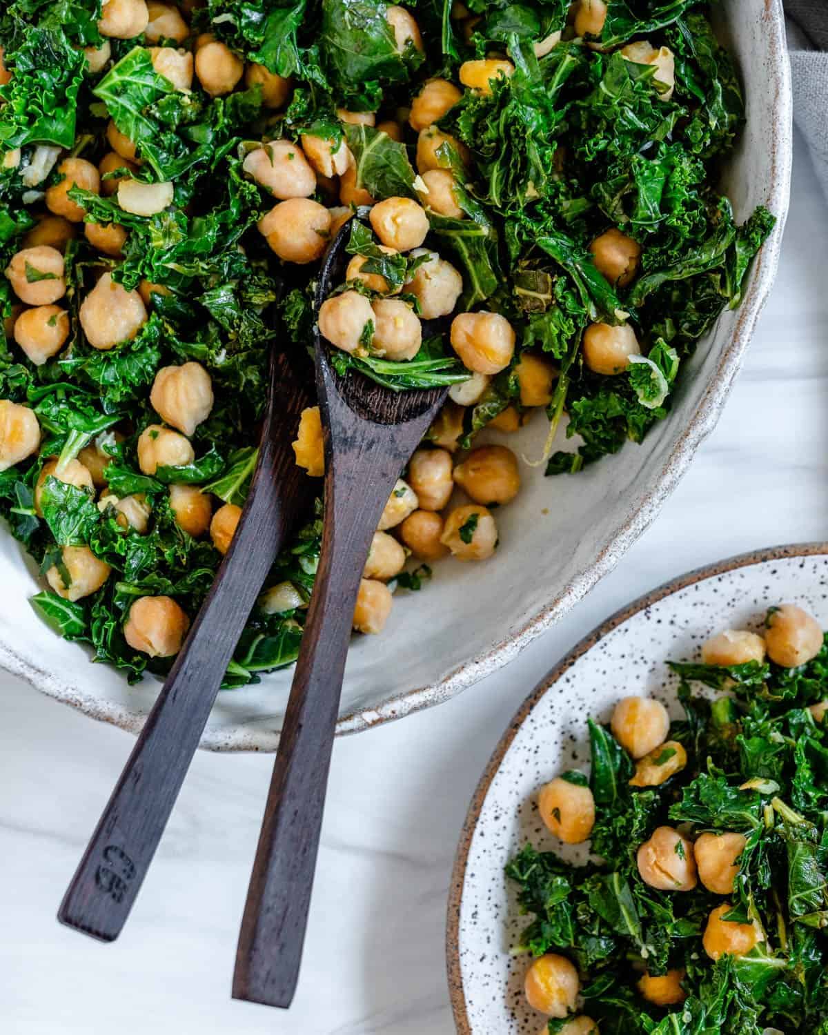 completed Garbanzo Bean Kale Salad plated on a platter and plate against a white background with serving utensils in the salad