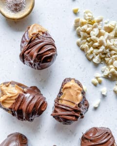 process of adding chocolate to peanut butter stuffed dates on white surface with nuts in the background