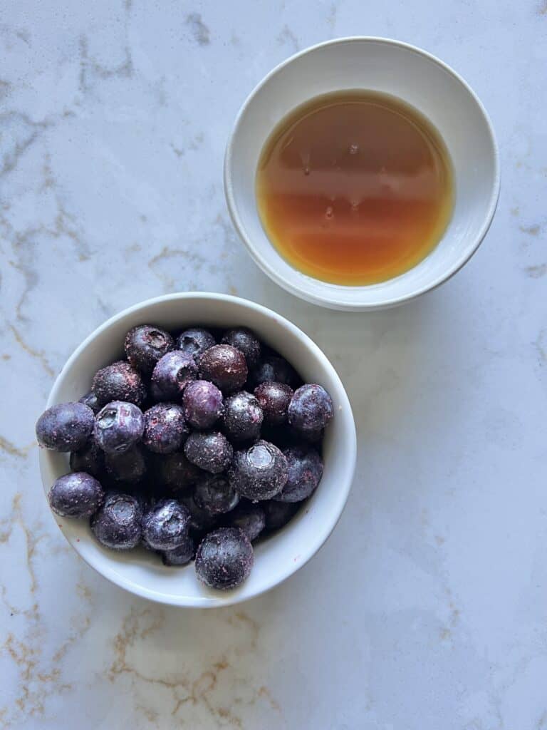 small white bowl of blueberries and small white bowl of maple syrup against white marble background