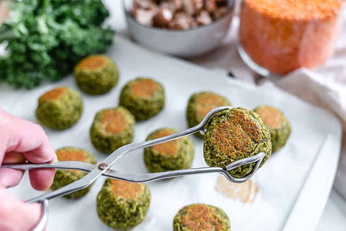 completed red lentil meatballs lined up and evenly spaced on a white surface with one being picked up with tongs