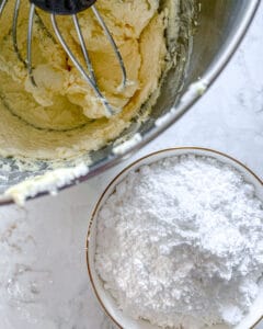 powdered sugar in a bowl alongside batter with whisk in the batter against white surface