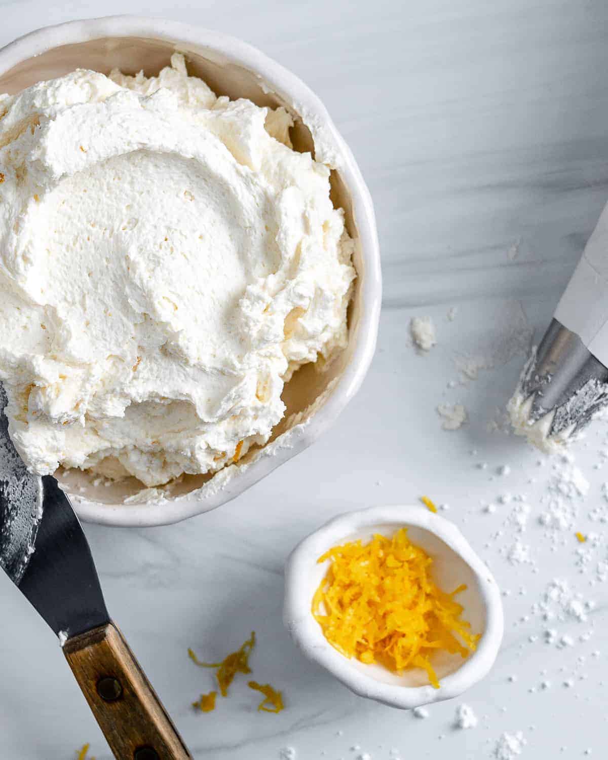 completed Lemon Buttercream Frosting in a white bowl against a white background with lemon zest in the background