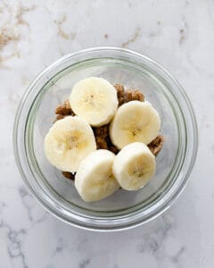 addition of banana slices on top of granola and whipped cream and banana mixture in clear jar against white marble background