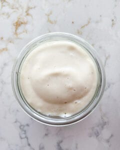 process of banana cream mixture on top of granola in a glass container against a white marble background