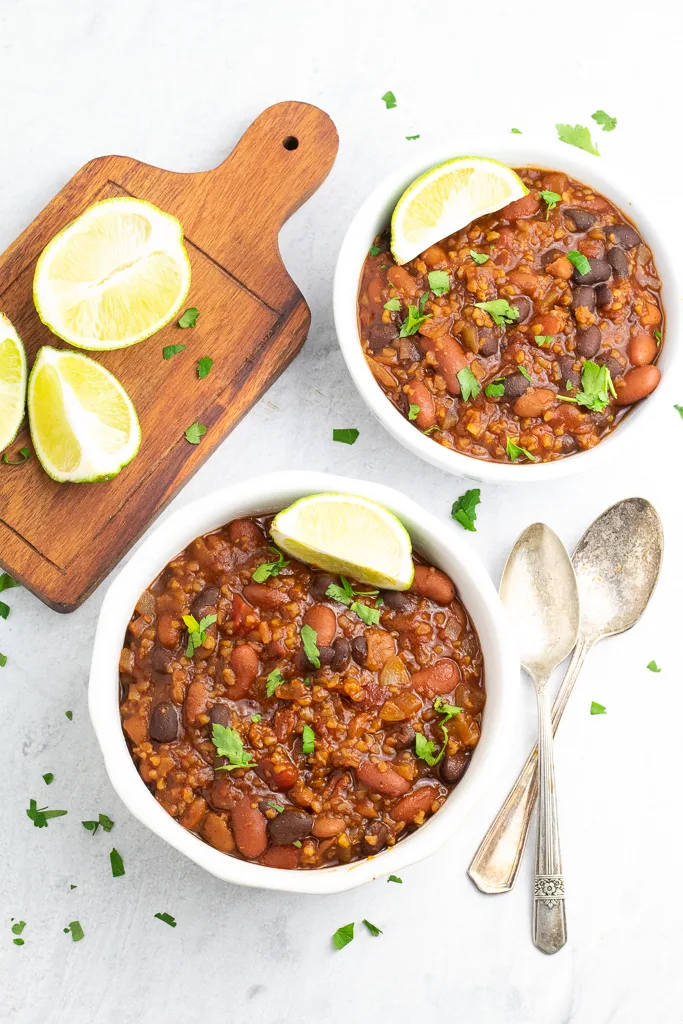 completed vegan chili in two white bowls with a cutting board with lemons and utensils against a white background