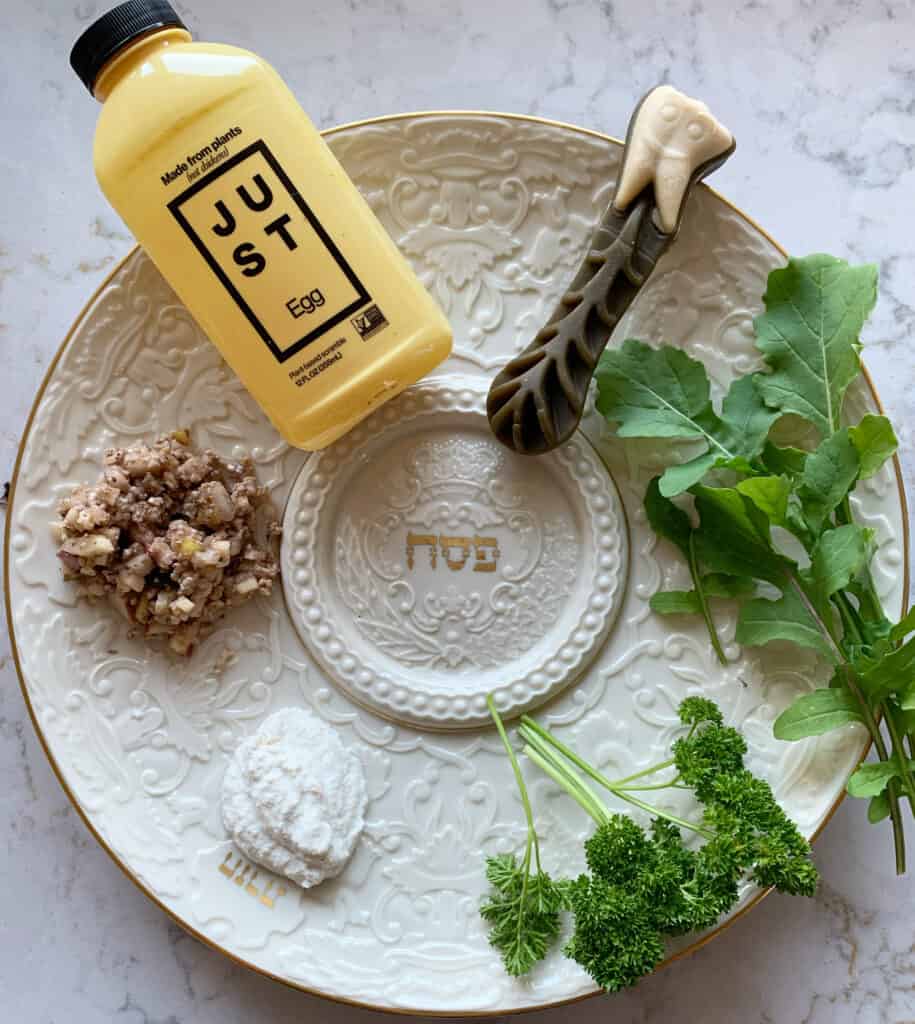 A white seder plate on a white marble table showing horseradish, parsley, arugula, charoset, a whimzee bone, and a Just Egg bottle.