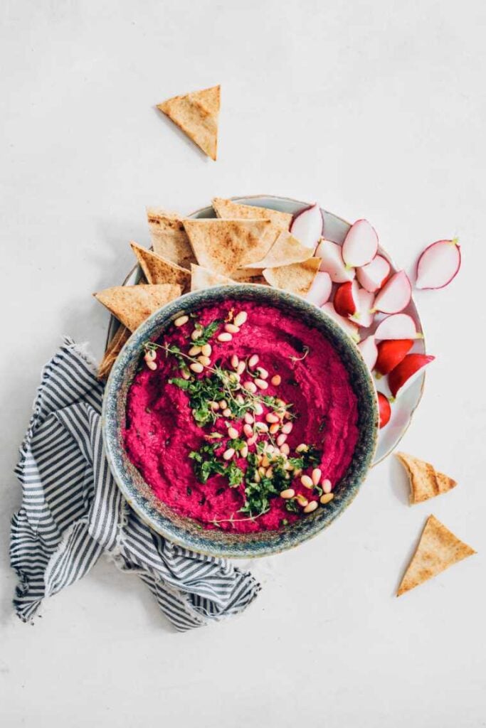 completed beet hummus in a white bowl on a plate with chips and veggies against a white background