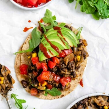 completed Vegan Black Bean Tacos [2 Ways] on a white surface with ingredients scattered in the background