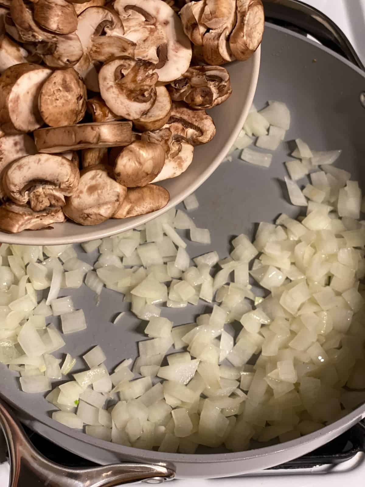 Sliced mushrooms being added to the cooked onion in a skillet.