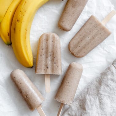 completed Healthy Banana Popsicles against a white surface alongside bananas