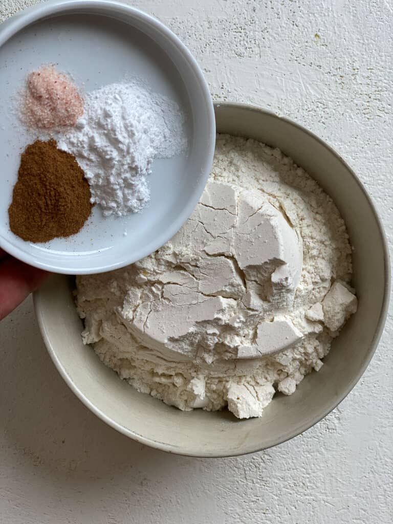 process of adding salt, baking powder, and cinnamon to bowl of vegan apple muffin mixture